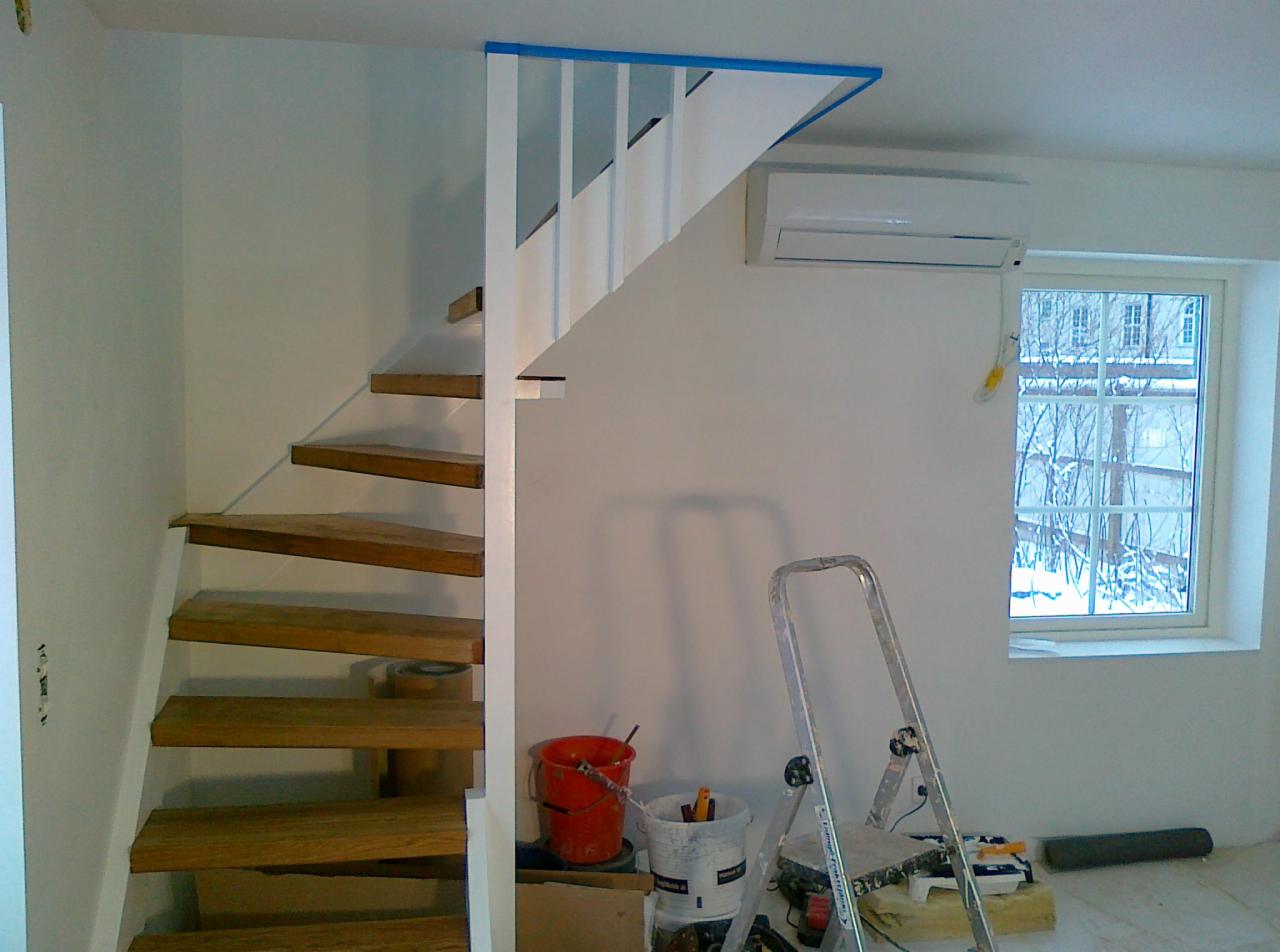 Drewniane schody spiralne z białymi barierkami, częściowo pomalowane, z niebieską taśmą malarską na górze, widoczne narzędzia malarskie, drabina i okno z widokiem na zimowy krajobraz.