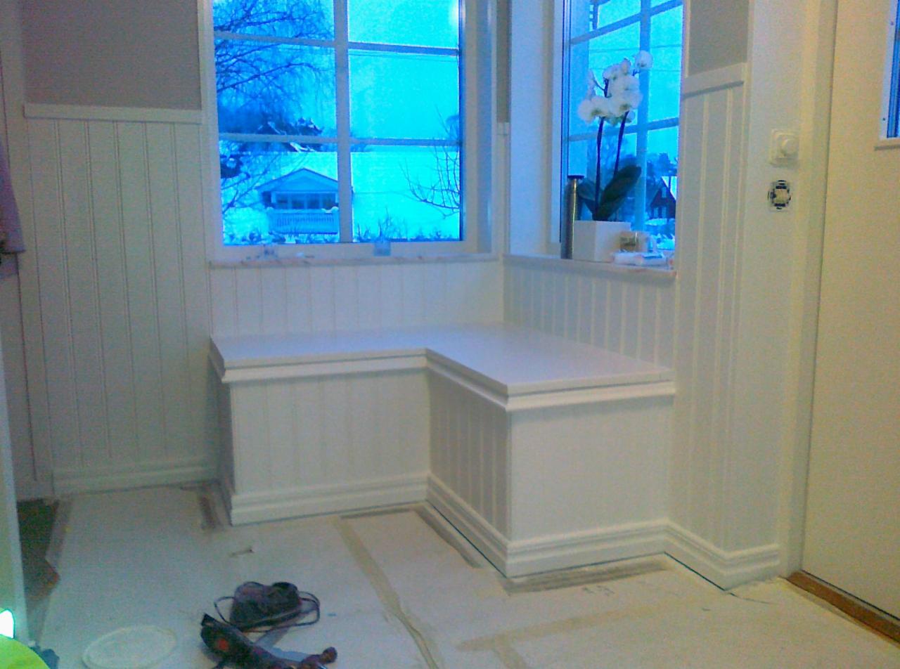 Biała, zabudowana ławka narożna z drewnianymi panelami ściennymi w pokoju z oknem, przez które widać zimowy krajobraz i dom, narzędzia leżą na podłodze zabezpieczonej folią.