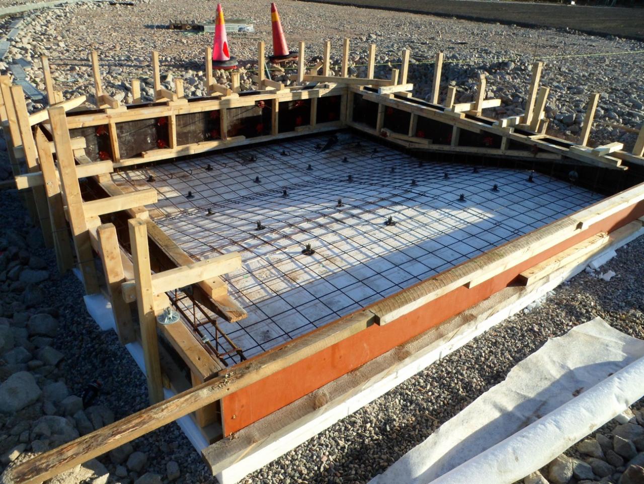 Szczegółowy widok szalunku zbrojeniowego na podłożu żwirowym, przygotowanego do wylania betonu, z widocznymi elementami drewnianymi, siatką zbrojeniową i dystansami, w tle dwa pomarańczowe pachołki...