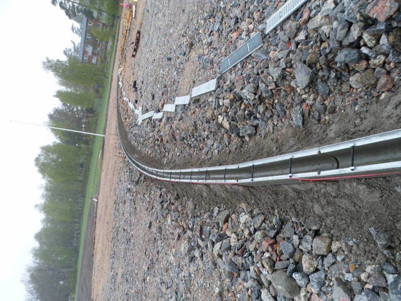 Długi, zakrzywiony drenaż liniowy z betonowym korytem i metalową kratką, ułożony w wykopie wypełnionym żwirem, widoczne elementy mocujące i czerwone przewody.