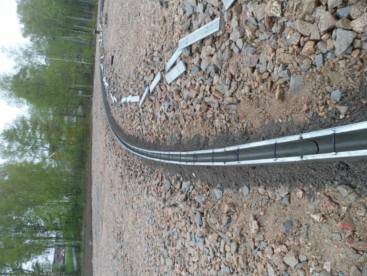 Zakrzywiony, metalowy kanał drenażowy, częściowo zasypany ziemią, ułożony na żwirowym podłożu wzdłuż granicy z drzewami.