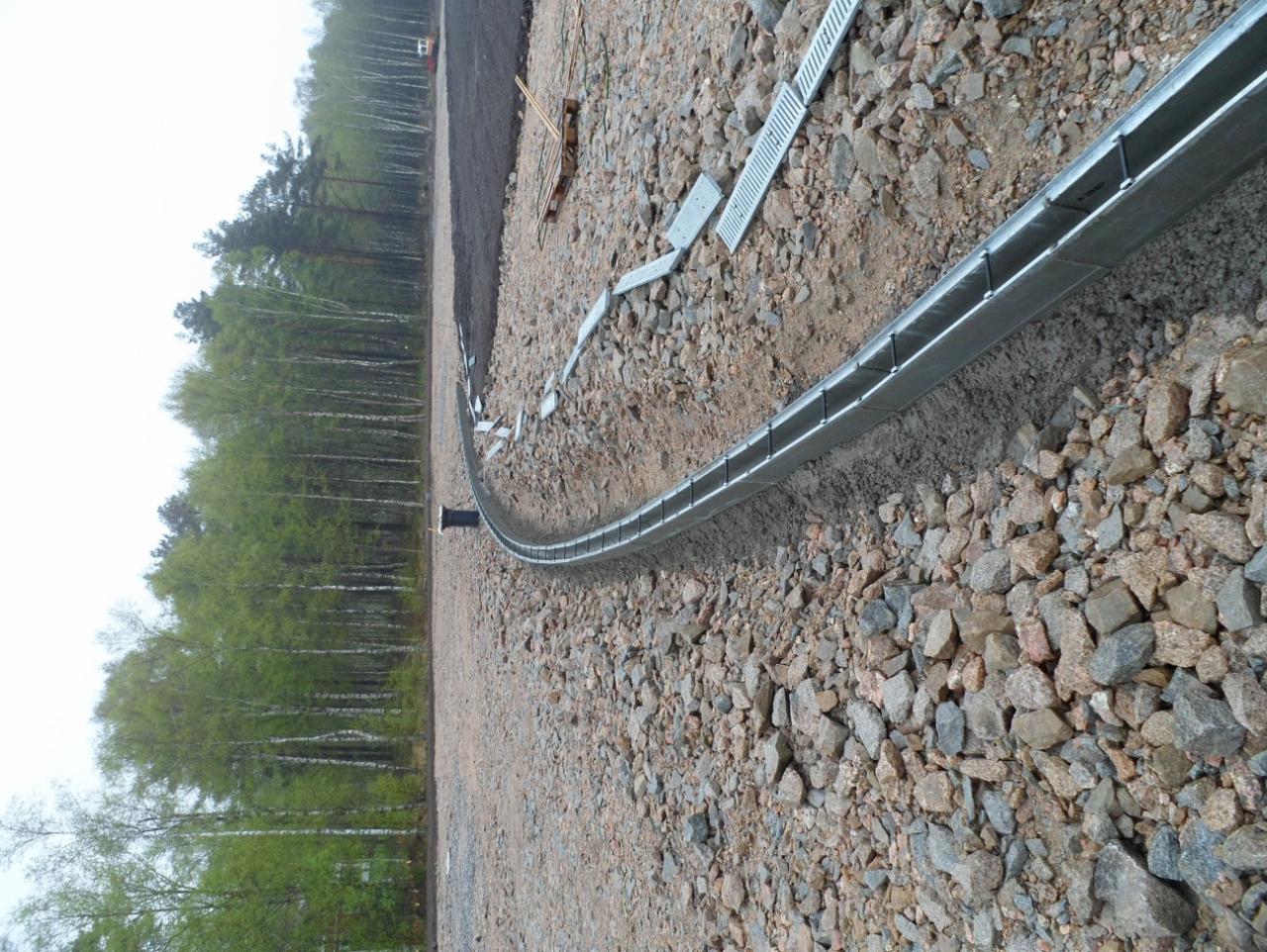 Instalacja drenażu liniowego z metalową rynną na żwirowym podłożu, widoczne elementy mocujące i fragmenty korytek, w tle las brzozowy i przygotowany teren pod budowę.
