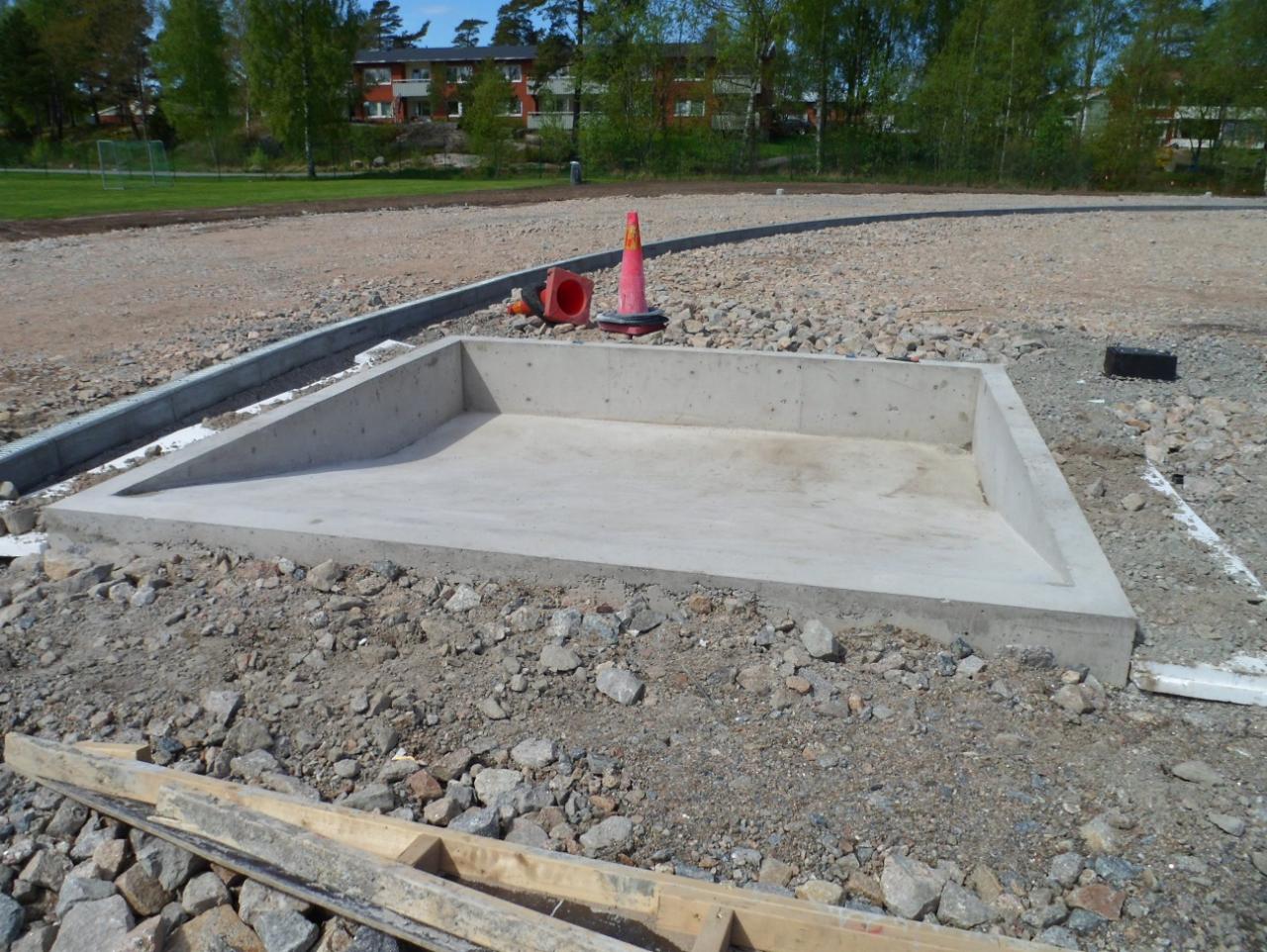 Betonowa podstawa w kształcie prostokąta, prawdopodobnie fundament pod budowlę, otoczona żwirem i gruzem. W tle widoczny trawnik, drzewa i budynek mieszkalny.