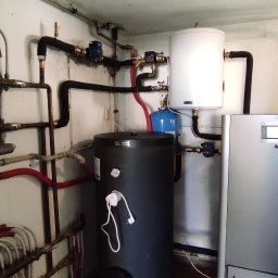 Oleksii Smal - Kotłownia z czarnym zasobnikiem ciepłej wody, białym zbiornikiem wyrównawczym i szarym piecem CO. Widoczne rury instalacji grzewczej i wodnej.