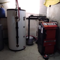 Oleksii Smal - Kotłownia z piecem CO Atmos, zbiornikami buforowymi i instalacją. Widoczne rury, zawory i okno z narzędziami. Surowe ściany z betonu komórkowego.