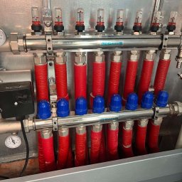 Oleksii Smal - Rozdzielacz ogrzewania podłogowego marki Gross z czerwonymi rurami i niebieskimi zaworami, zamontowany w metalowej szafce, widok z bliska.