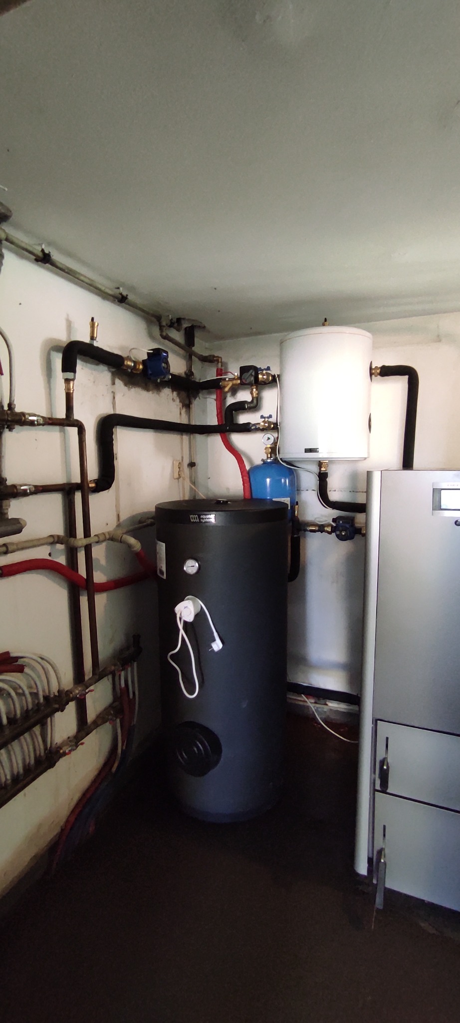 Kotłownia z czarnym zasobnikiem ciepłej wody, białym zbiornikiem wyrównawczym i szarym piecem CO. Widoczne rury instalacji grzewczej i wodnej.