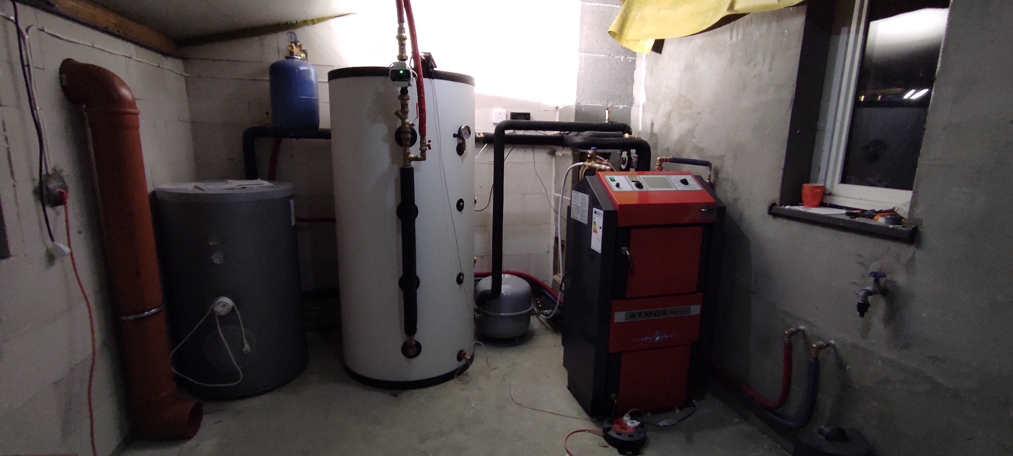 Kotłownia z piecem CO Atmos, zbiornikami buforowymi i instalacją. Widoczne rury, zawory i okno z narzędziami. Surowe ściany z betonu komórkowego.