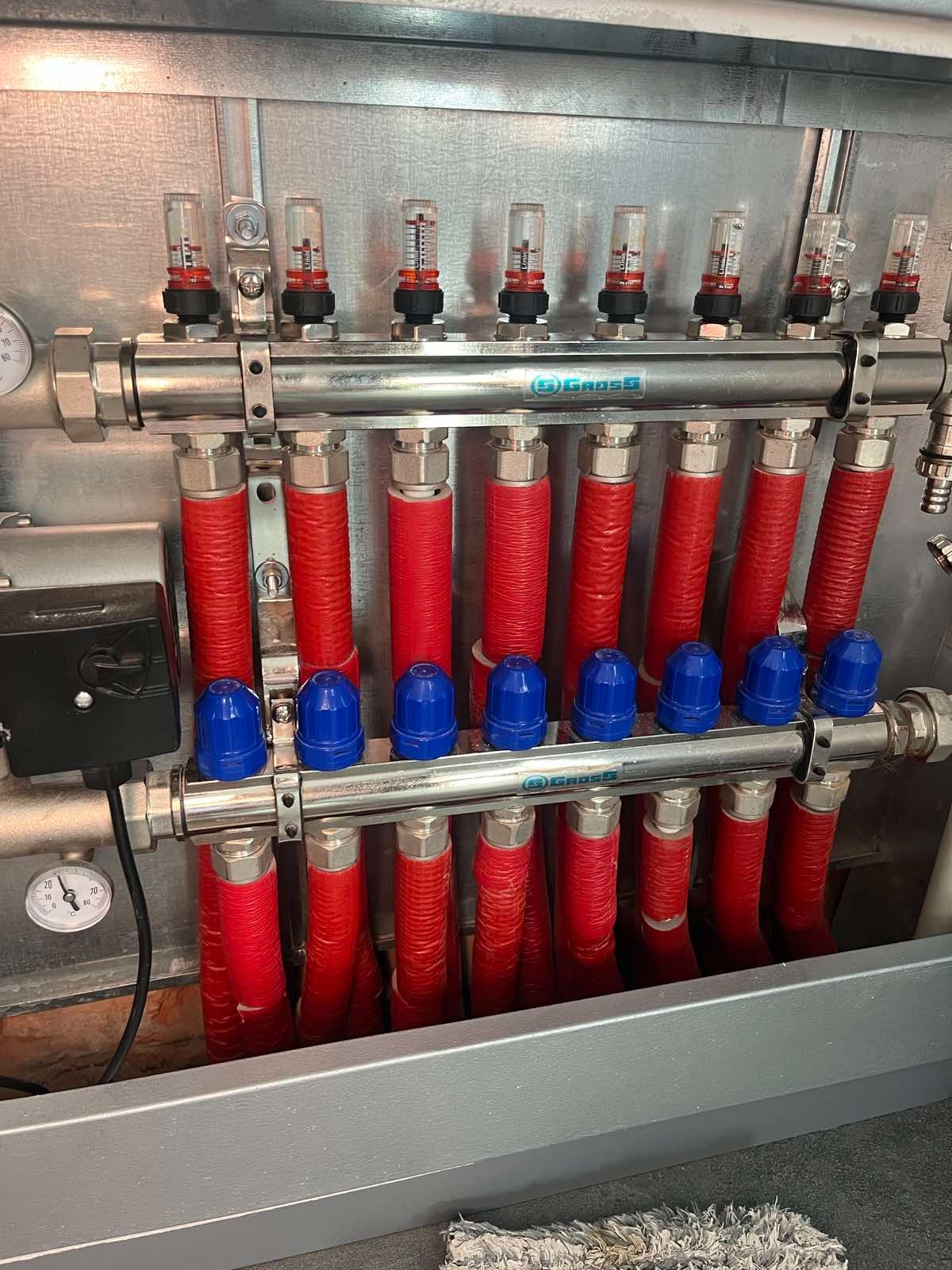 Rozdzielacz ogrzewania podłogowego marki Gross z czerwonymi rurami i niebieskimi zaworami, zamontowany w metalowej szafce, widok z bliska.