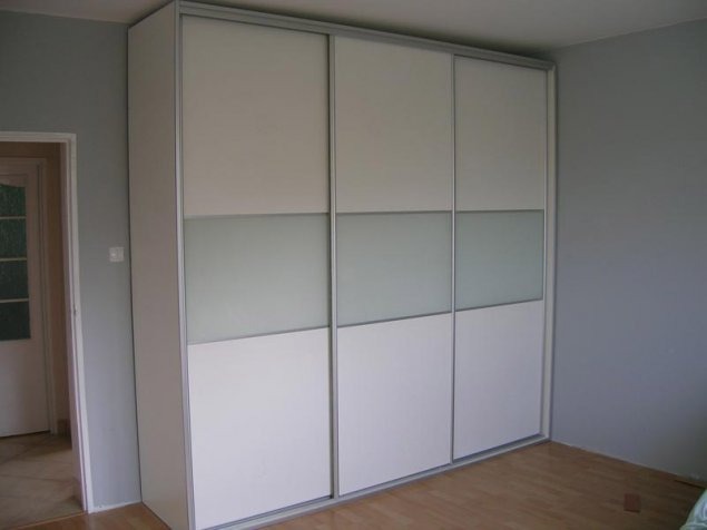 Biała szafa przesuwna z trzema drzwiami, z poziomym pasem z matowego szkła, na tle szarej ściany i drewnianej podłogi. Widok z boku, oświetlenie dzienne.