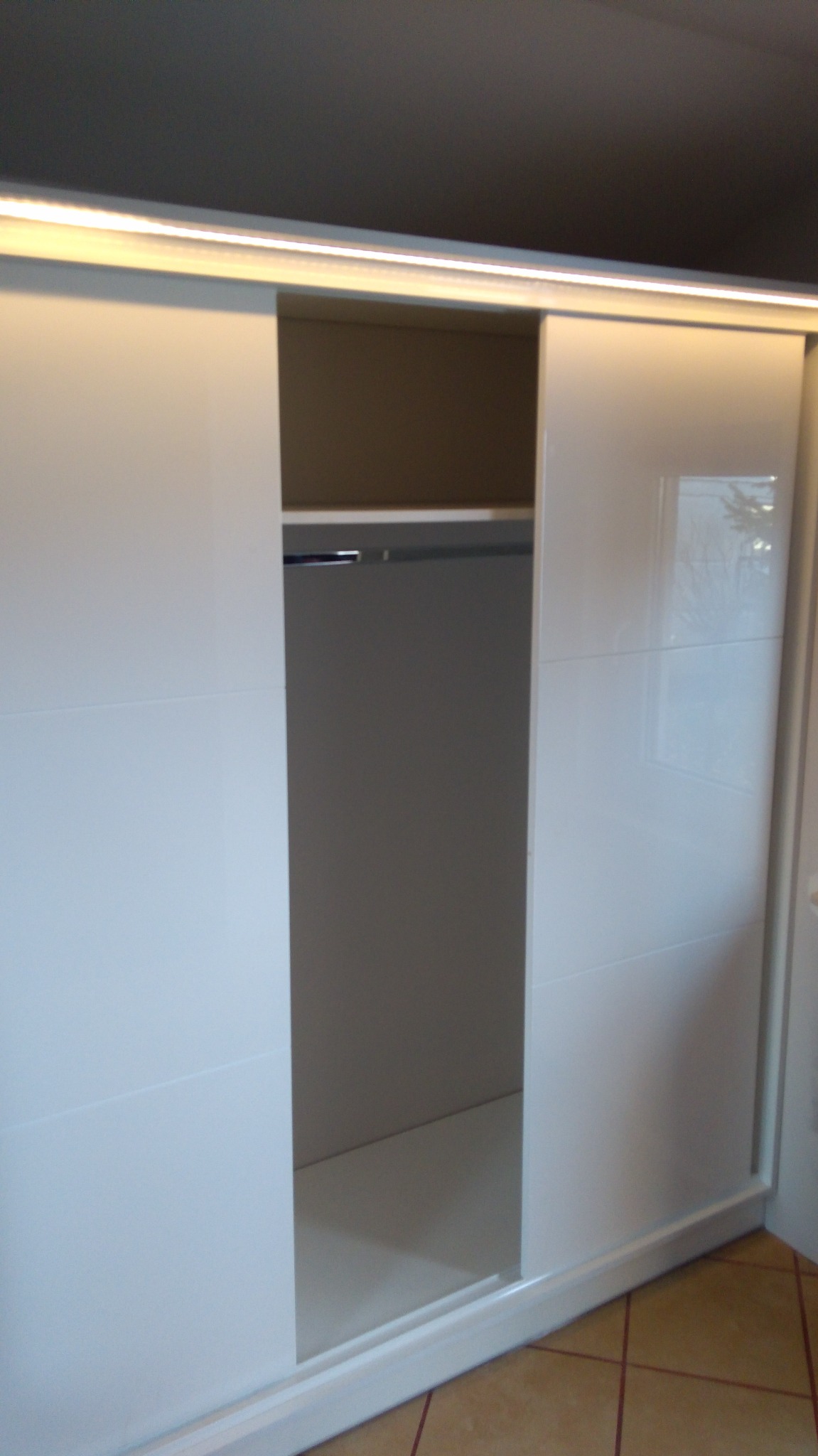 Biała szafa przesuwna z oświetleniem LED u góry, częściowo otwarta, ukazująca wnętrze z półką i drążkiem na ubrania. Widoczna podłoga z płytek.