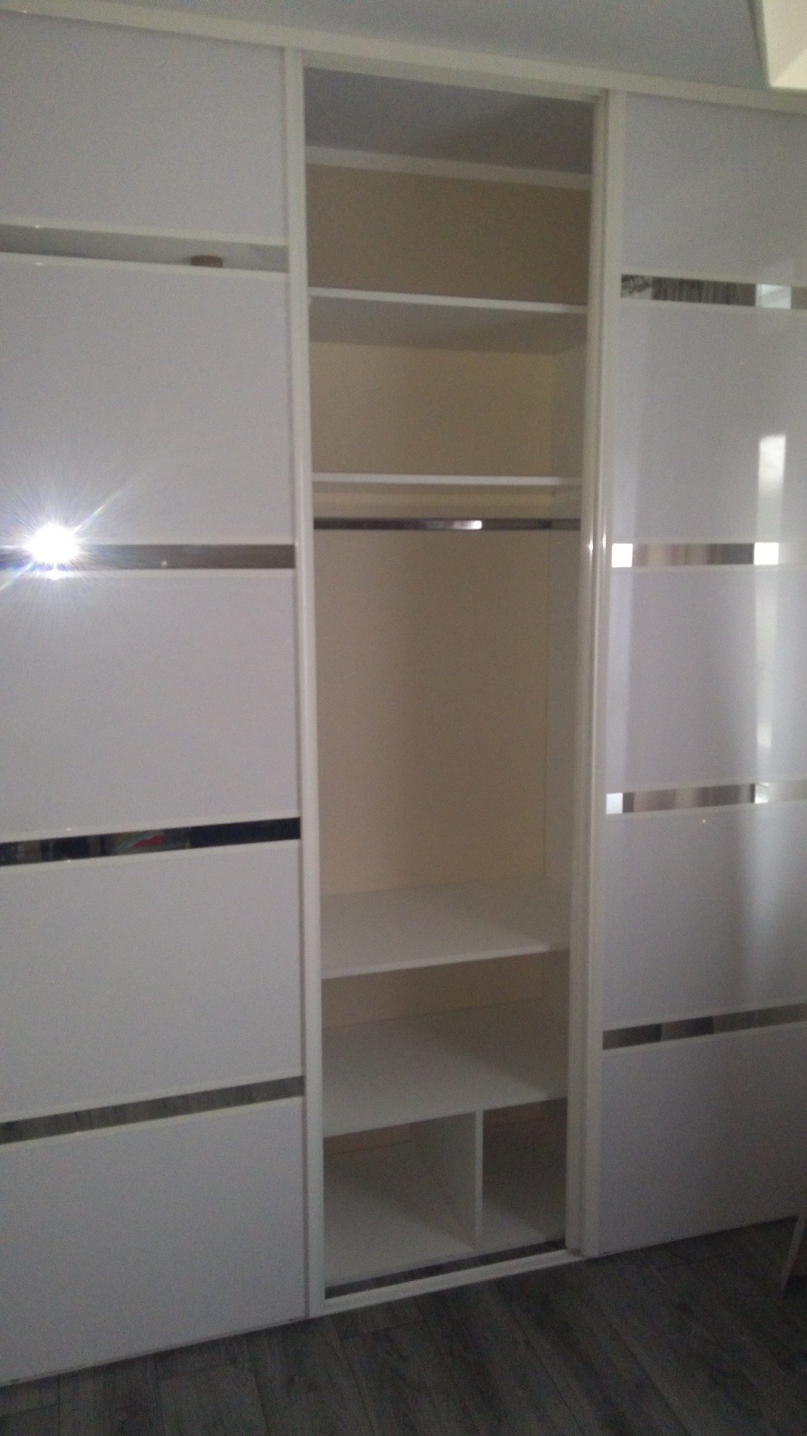 Biała szafa przesuwna z lustrzanymi wstawkami, widoczne półki i drążek na ubrania. Nowoczesny design, minimalistyczne wykończenie.