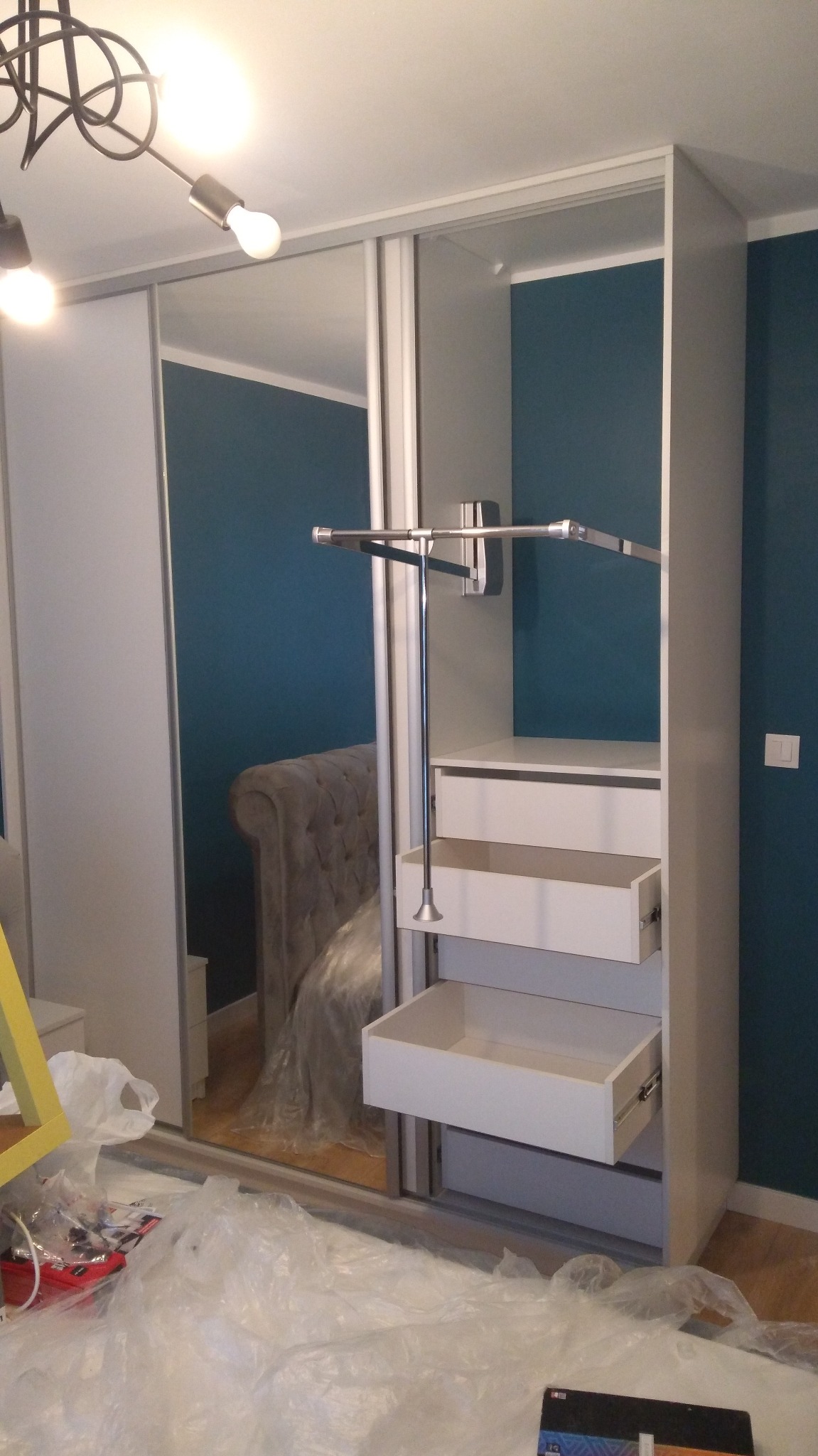 Biała szafa przesuwna z lustrem i wysuwanymi szufladami, widoczny drążek na ubrania, w tle łóżko z wezgłowiem. Podłoga zabezpieczona folią malarską.