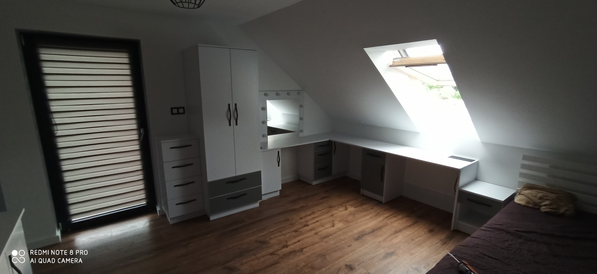 Białe meble na wymiar w pokoju na poddaszu: szafa, toaletka z oświetleniem i biurko pod oknem dachowym. Parkiet na podłodze.