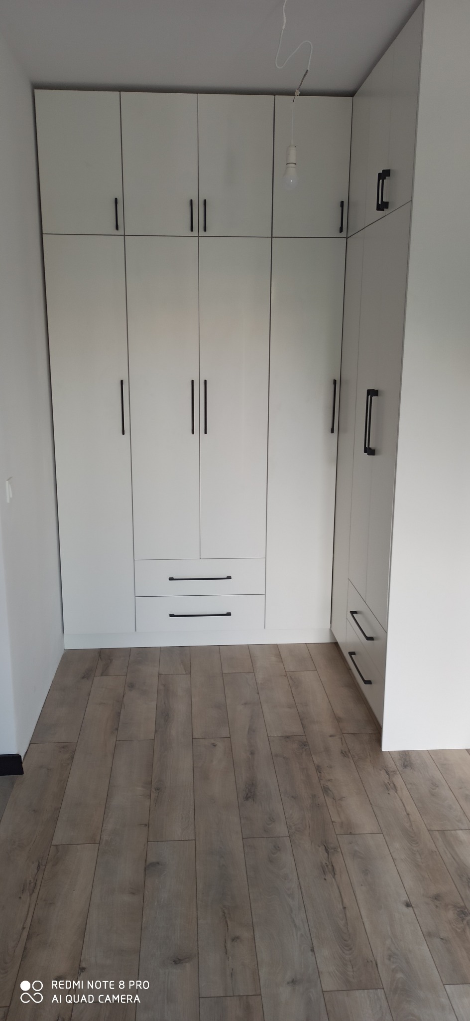 Biała zabudowa szafy z szufladami i czarnymi uchwytami, widok na jasną podłogę z paneli. Minimalistyczny design, proste linie, sufit z wiszącą żarówką.