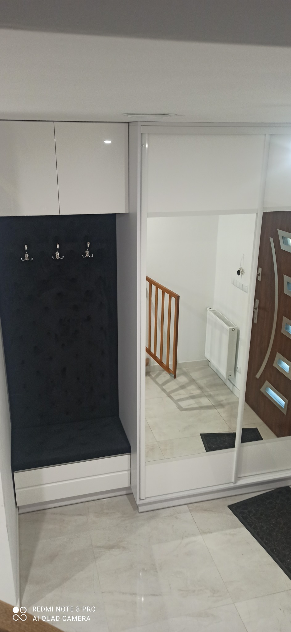 Biała szafa wnękowa z lustrem i siedziskiem z wieszakami w przedpokoju. Widoczne odbicie schodów i drzwi wejściowych. Nowoczesny design, jasne wnętrze.