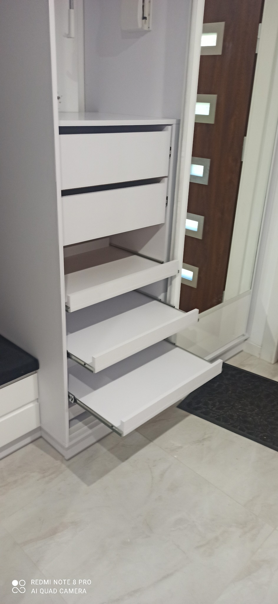 Biała szafa z wysuwanymi półkami i szufladami, widok z boku, wnętrze szafy, drzwi z elementami dekoracyjnymi, dywanik pod drzwiami.