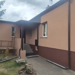 Wykończona elewacja domu jednorodzinnego w odcieniach beżu i brązu z widocznymi schodami wejściowymi, Poznań. Ujęcie z poziomu gruntu, w tle zieleń.