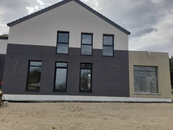 Dwukolorowa elewacja budynku w trakcie prac wykończeniowych; część ściany w szarym tynku, a druga w beżowym, okna zabezpieczone folią.
