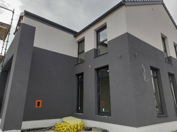 Dwukolorowa elewacja domu po tynkowaniu z szarym dołem i białym górnym pasem. Widoczne okna w ciemnych ramach i fragment rusztowania po lewej stronie.
