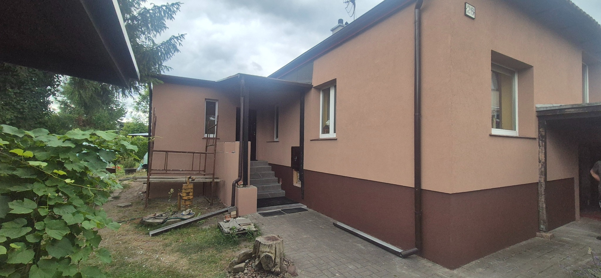 Wykończona elewacja domu jednorodzinnego w odcieniach beżu i brązu z widocznymi schodami wejściowymi, Poznań. Ujęcie z poziomu gruntu, w tle zieleń.