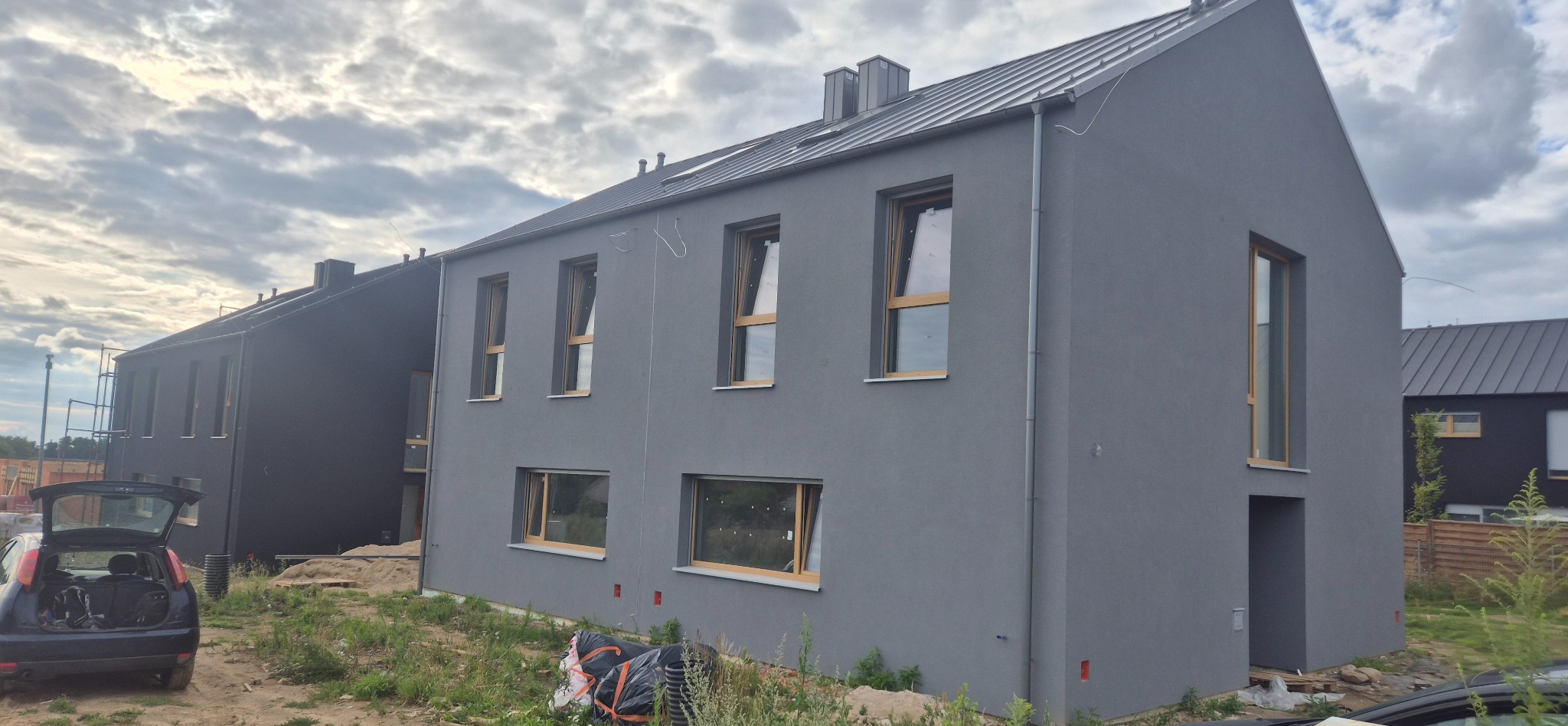 Szare, dwukondygnacyjne domy w zabudowie szeregowej z drewnianymi oknami i metalowymi dachami, w trakcie wykończenia elewacji. Widoczne worki z materiałami budowlanymi.