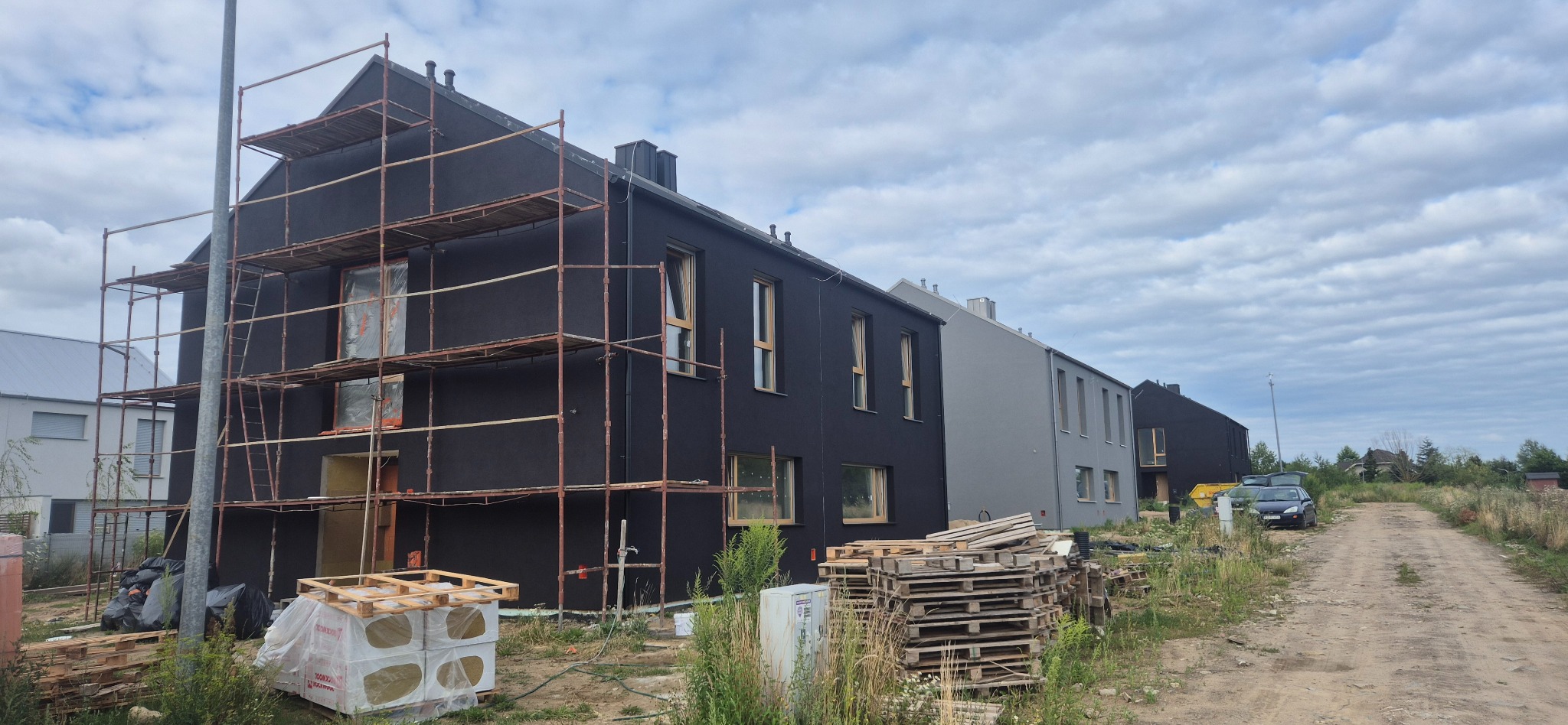 Budynek w trakcie prac elewacyjnych: rusztowania, częściowo otynkowana, czarna elewacja, okna z drewnianą stolarką. Na pierwszym planie palety.