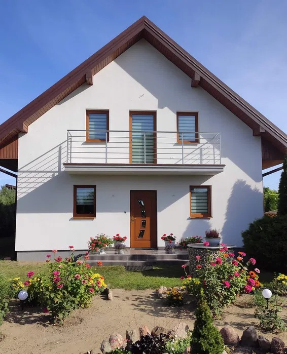 Biały dom jednorodzinny z brązowym dachem i drewnianymi oknami, balkon z metalową barierką, kwiaty przed wejściem, zadbany ogród, słoneczny dzień.