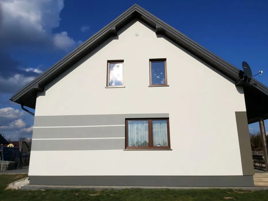 Elewacja domu jednorodzinnego z widocznym szarym pasem dekoracyjnym, okna z brązowymi ramami, dachówka w kolorze grafitu i fragment ogrodu.