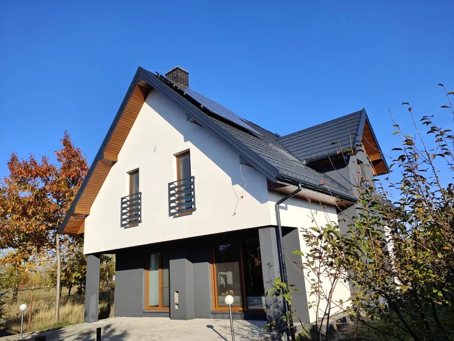 Dwukondygnacyjny dom z białą elewacją i szarymi akcentami, widoczny fragment dachu z panelami słonecznymi, okna z balustradami, jesienne drzewo w tle.