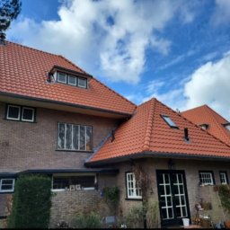 Kamil Soltysinski - Ceglasty dach z lukarnami na dwukondygnacyjnym domu z cegły, widok z poziomu ogrodu w słoneczny dzień. Detale: czarne obramowania okien i drzwi.