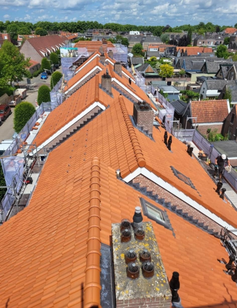 Pomarańczowy dach z dachówki ceramicznej na szeregu domów, widok z góry, z kominami i oknami dachowymi. Prace dekarskie w toku, widoczne rusztowania i pracownik.