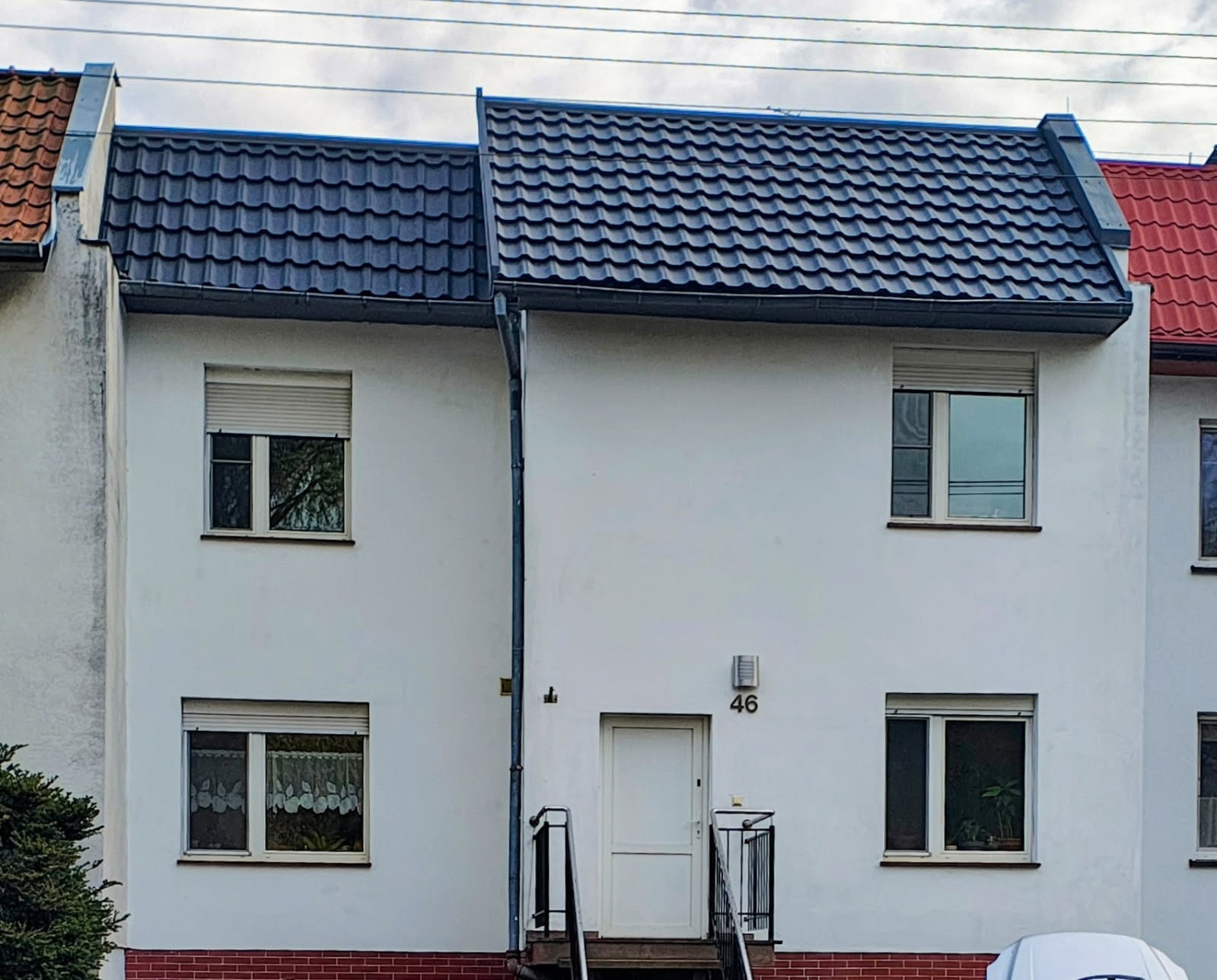 Bliźniak z ciemną dachówką ceramiczną, białą elewacją i numerem 46 nad wejściem. Widoczne rynny i okna z roletami. Fragment dachu sąsiedniego budynku z czerwoną dachówką.