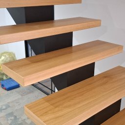 ATELIER.TOM - Nowoczesne schody z drewnianymi stopniami i czarną, metalową konstrukcją nośną. Widok z dołu, ukazujący detale wykończenia i minimalistyczny design. Pod schodami leży pies.