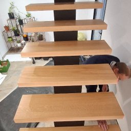 ATELIER.TOM - Montaż drewnianych schodów wspornikowych z czarną konstrukcją w Radłowie. Widok z dołu, osoba montuje stopnie. Minimalistyczny design, jasne drewno.