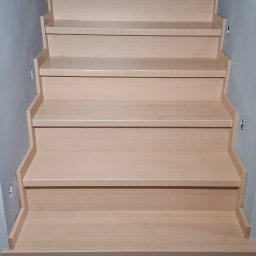 ATELIER.TOM - Jasne, drewniane schody z podświetleniem LED na ścianie, widziane z dołu. Widoczne narzędzia i materiały budowlane sugerują etap wykończeniowy.