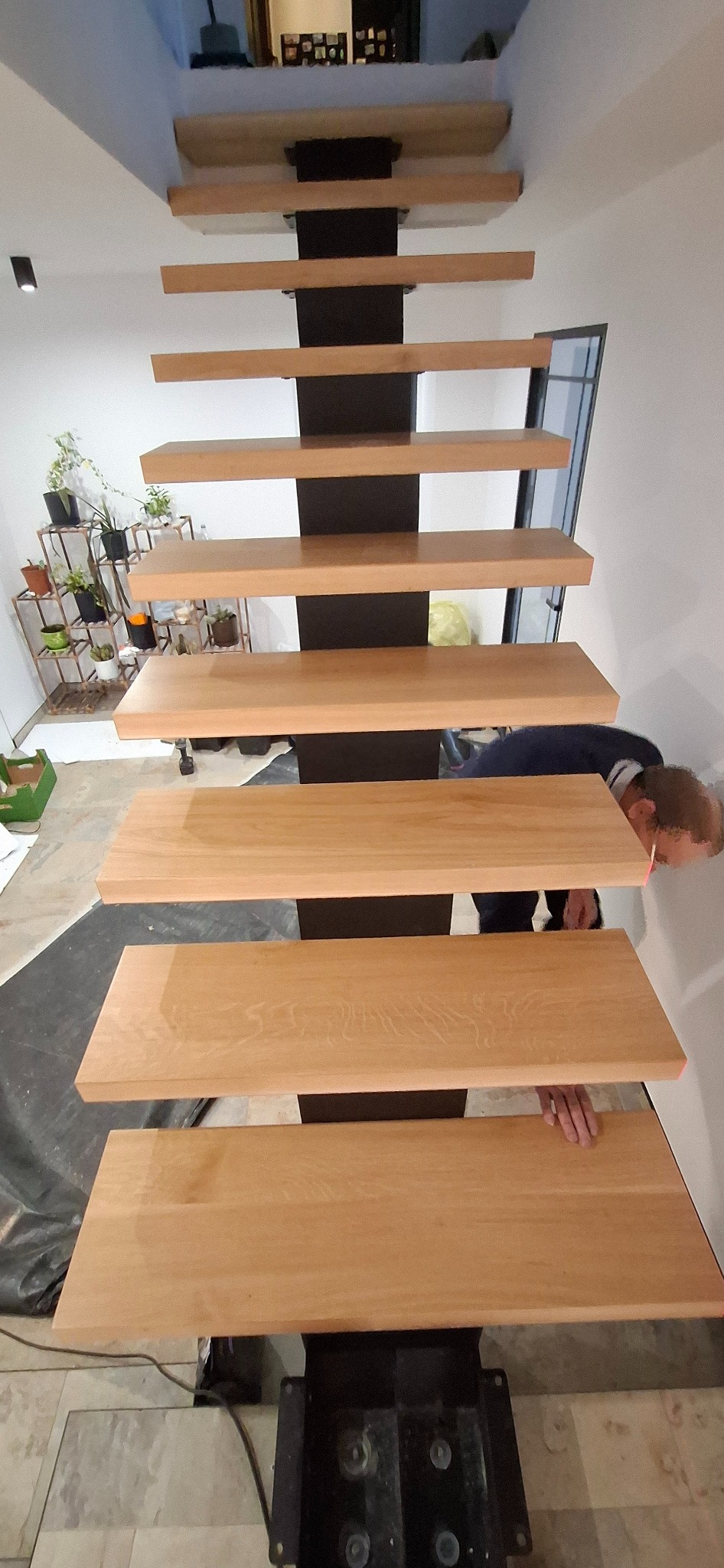 Montaż drewnianych schodów wspornikowych z czarną konstrukcją w Radłowie. Widok z dołu, osoba montuje stopnie. Minimalistyczny design, jasne drewno.