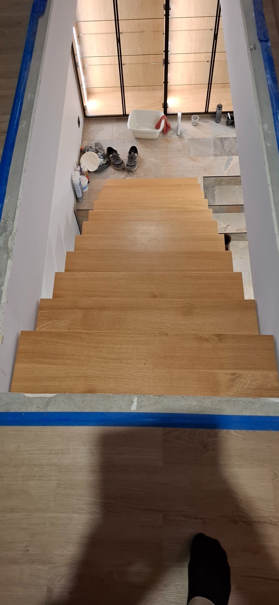 Jasne, drewniane schody w trakcie montażu, widok z góry. Zabezpieczone niebieską taśmą krawędzie, w tle widoczne narzędzia i obuwie.