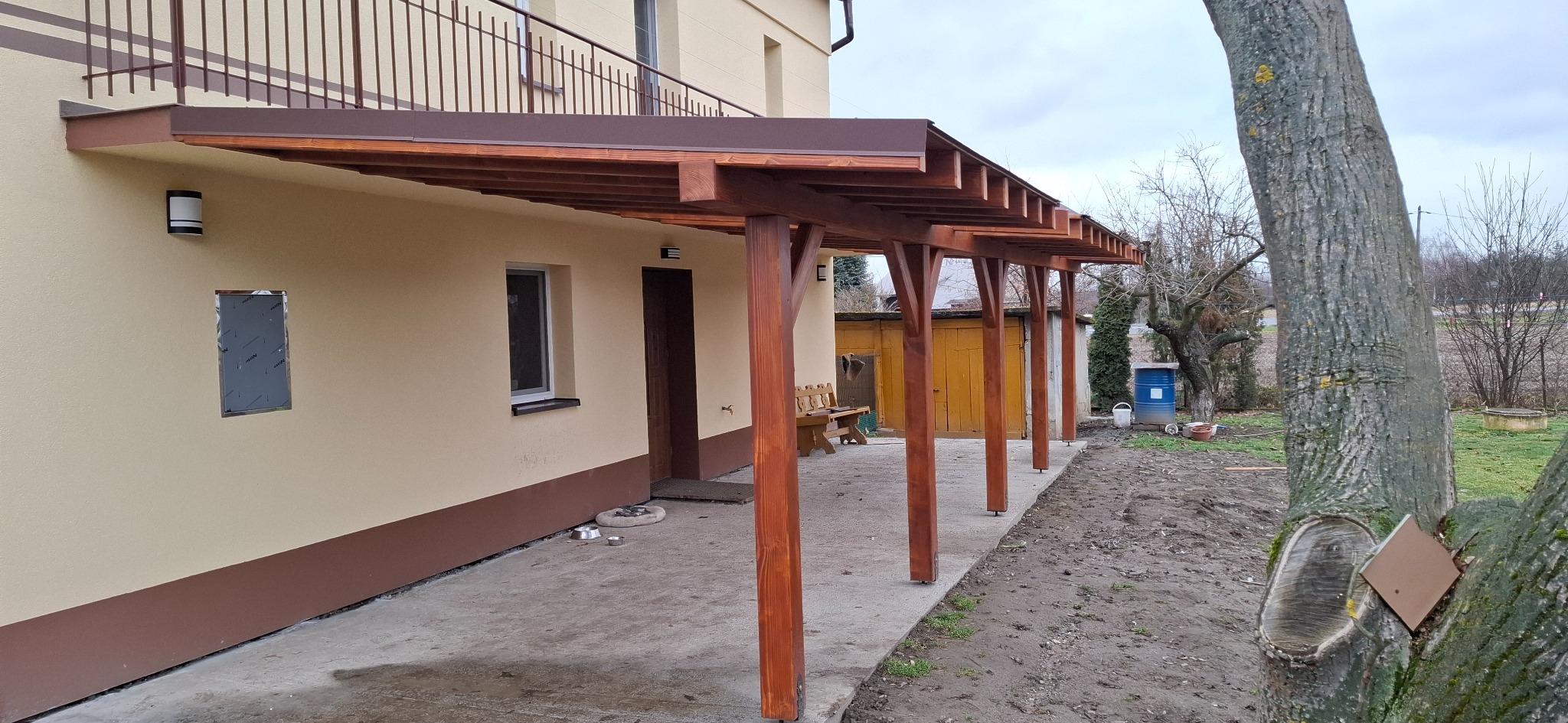 Drewniana pergola z brązowym zadaszeniem, przylegająca do beżowego budynku z brązowym cokołem. Widoczne słupy pergoli, okno, drzwi i fragment drzewa.