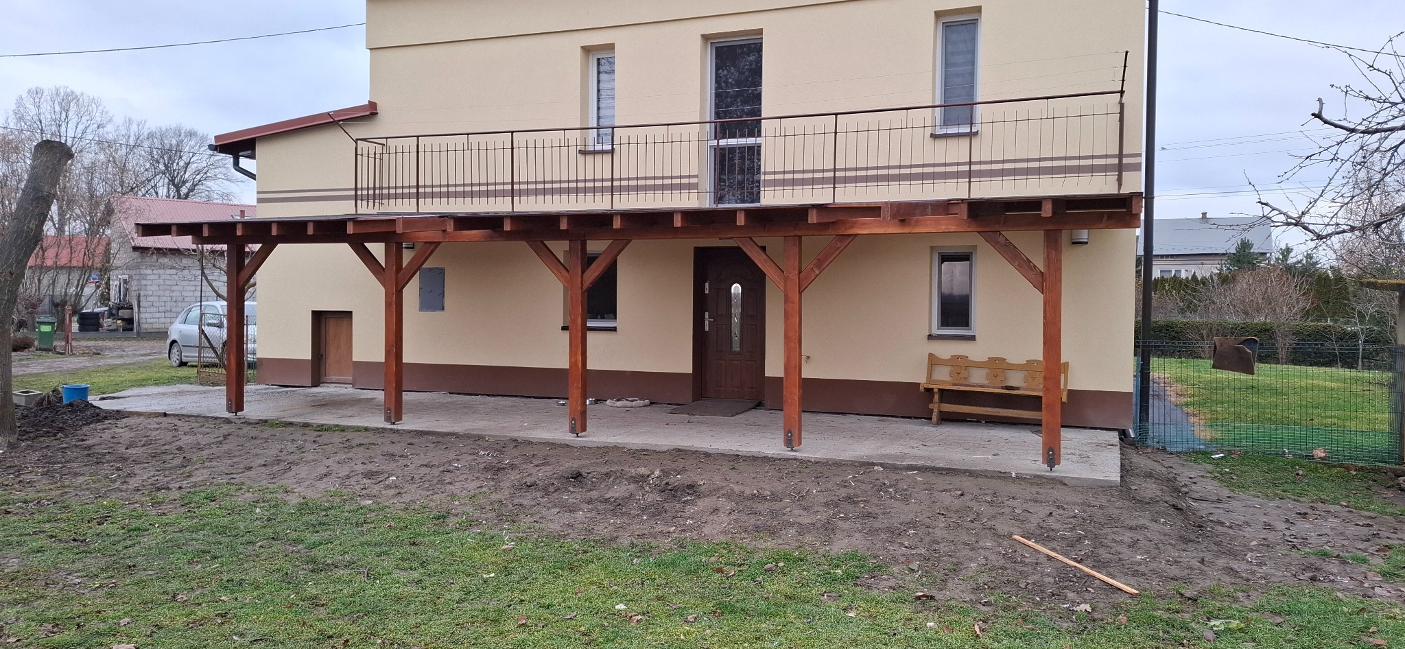 Drewniana pergola przy domu z balustradą na górze. Konstrukcja wsparta na solidnych słupach, z widocznym zadaszeniem. Ujęcie z zewnątrz, w tle budynek i zieleń.
