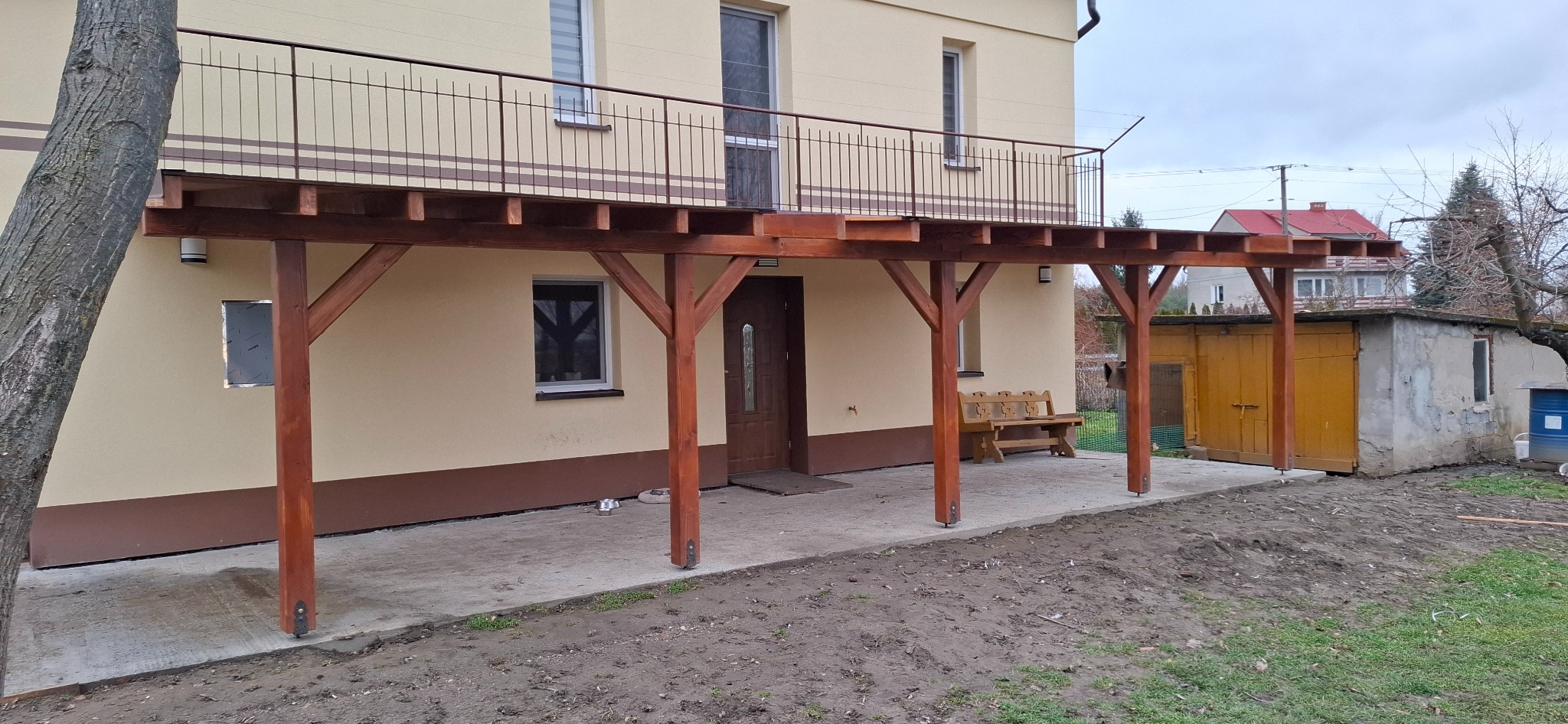 Drewniana pergola przy domu z balkonem na piętrze. Konstrukcja z belek w kolorze brązowym, podtrzymywana przez słupy. Widoczny fragment elewacji i otoczenie.