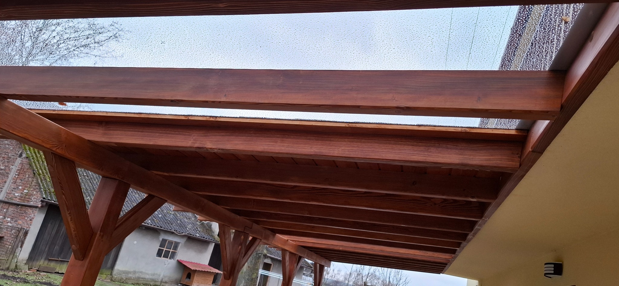 Drewniana pergola z przezroczystym zadaszeniem, widok od dołu. Konstrukcja z belek w ciepłym odcieniu brązu, w tle budynki i drzewa. Na zadaszeniu widoczne krople deszczu.