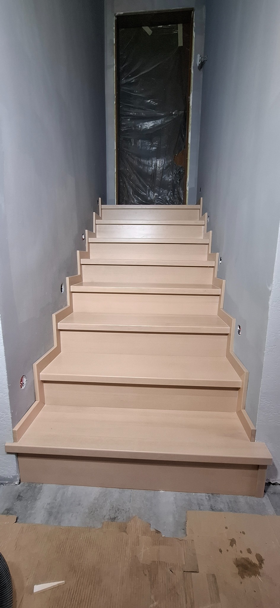 Jasne, drewniane schody z oświetleniem punktowym w szarej klatce schodowej. Drzwi zabezpieczone folią. Widok z dołu na górę.