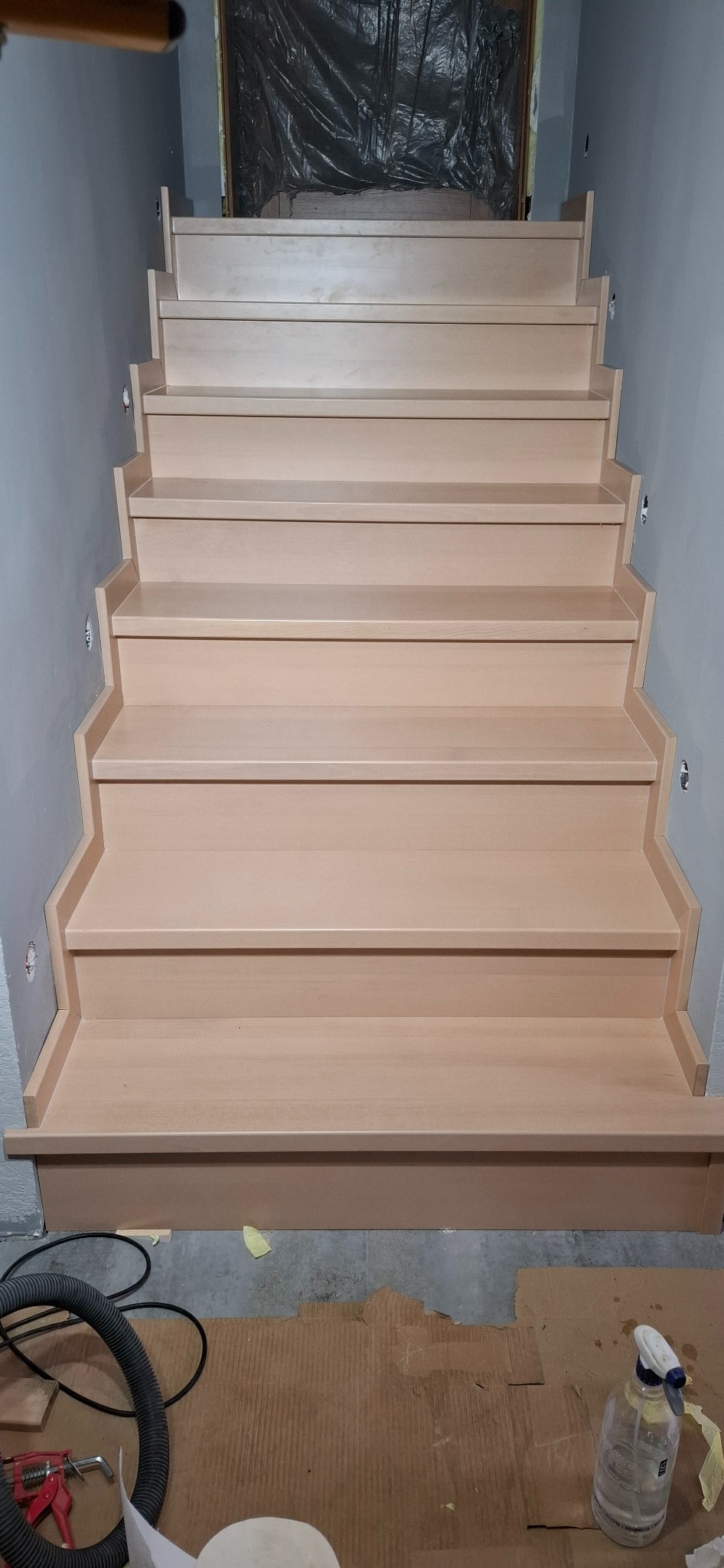 Jasne, drewniane schody z podświetleniem LED na ścianie, widziane z dołu. Widoczne narzędzia i materiały budowlane sugerują etap wykończeniowy.