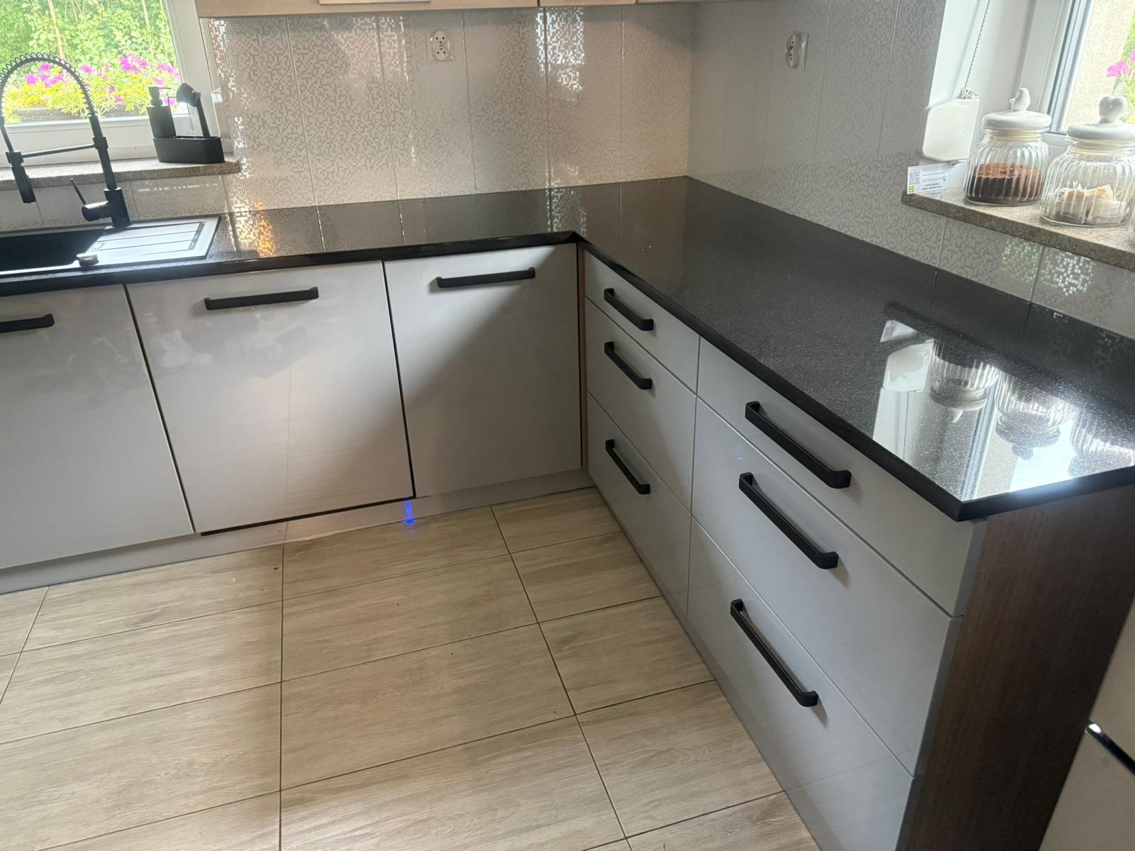 Nowoczesna kuchnia z białymi szafkami i czarnym blatem, minimalistyczne uchwyty, płytki imitujące drewno na podłodze, widok z niskiego kąta.