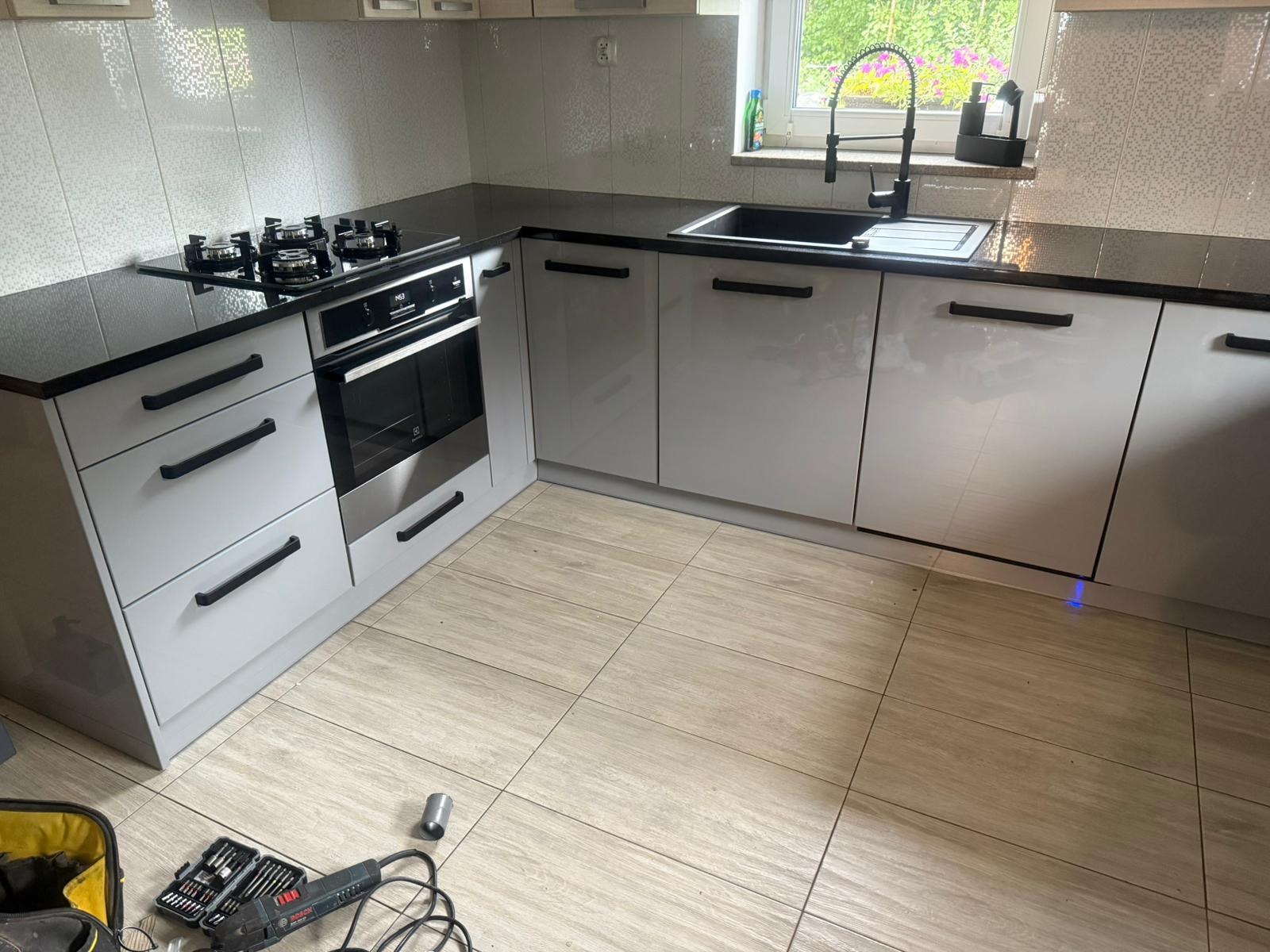 Jasnoszara kuchnia z czarnym blatem i sprzętem AGD. Widoczne narzędzia na podłodze sugerują świeży montaż.