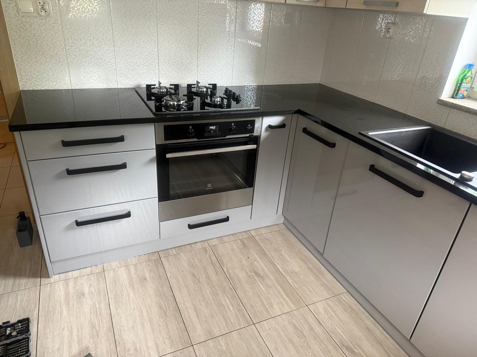 Nowoczesna kuchnia na wymiar z szarymi, lakierowanymi frontami, czarnym blatem i sprzętem AGD. Widoczna płyta gazowa i piekarnik. Jasne płytki na ścianie i podłodze.