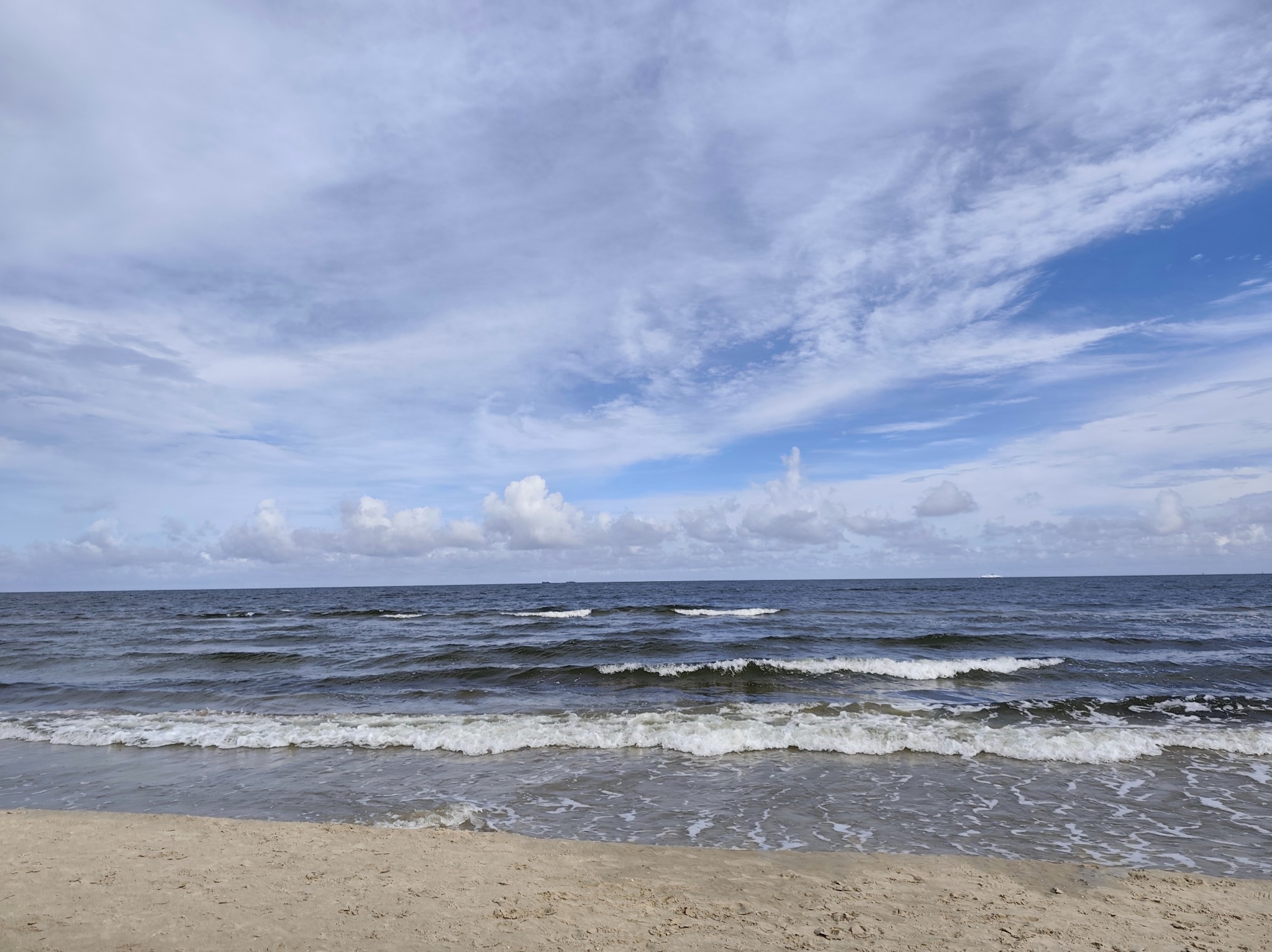 Spokojne morze z falami i piaszczystą plażą pod pochmurnym niebem. Horyzont widoczny w oddali, delikatne fale rozbijają się o brzeg.