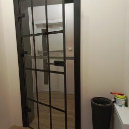 DAGMA - 29 lat doświadczenia w oknach i drzwiach - Częściowo otwarte, szklane drzwi wewnętrzne z czarną ramą, podzielone na kwadraty. Widoczna podłoga z paneli i białe ściany. Obok drzwi stoi kosz na śmieci i farba.