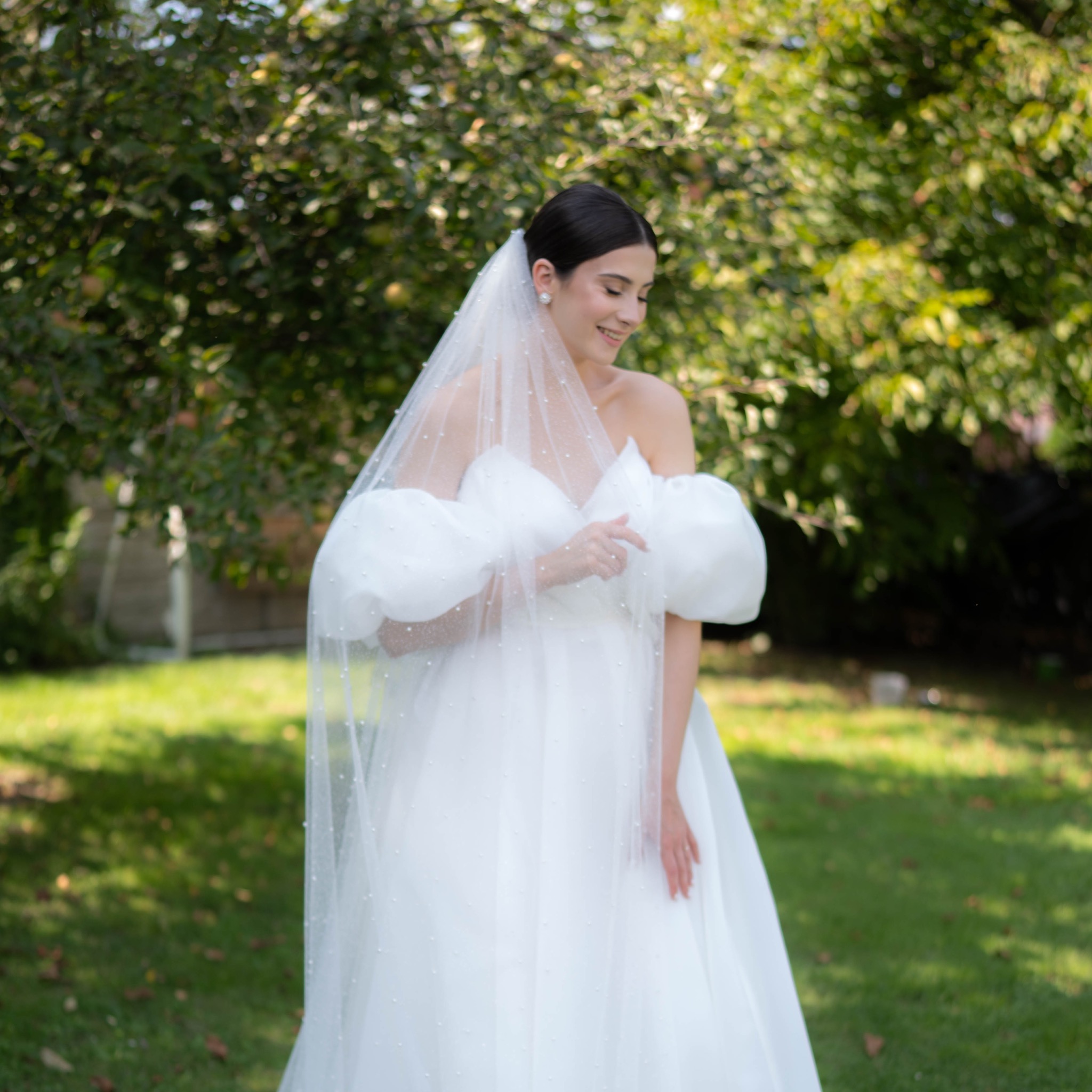 Uśmiechnięta panna młoda w białej sukni z bufiastymi rękawami i welonem w kropki pozuje na tle zieleni w plenerze. Delikatny makijaż, naturalne światło.